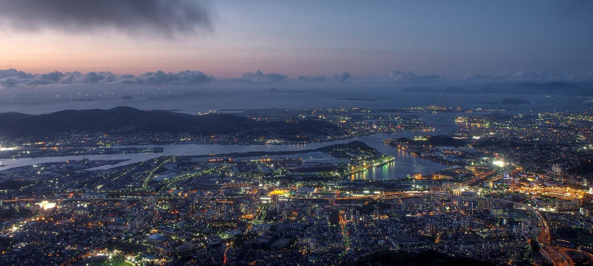 Fukuoka skyline at night, viewed from a mountain top. Bright city lights illuminate the urban landscape.