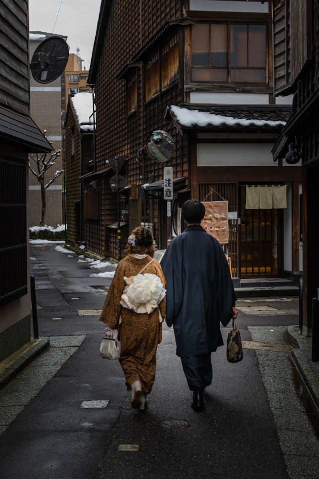 A couple dressed in traditional Japanese attire walks down a narrow, snow-covered street between wooden buildings.