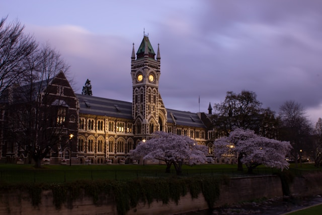 A clock tower standing tall next to a tree in front of a grand building.